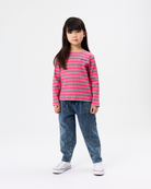 A young girl stands against a plain white studio background wearing a pink and light blue horizontally striped long-sleeve knit top, blue jeans, and white sneakers.