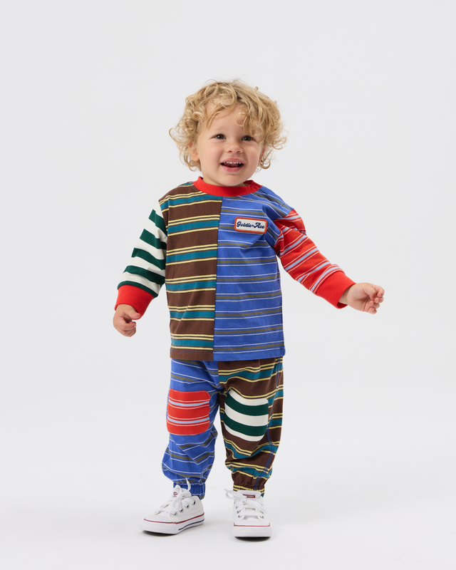 A toddler stands against a light gray studio background wearing a colorful patchwork long-sleeve pajama top and matching striped pants, with white sneakers.