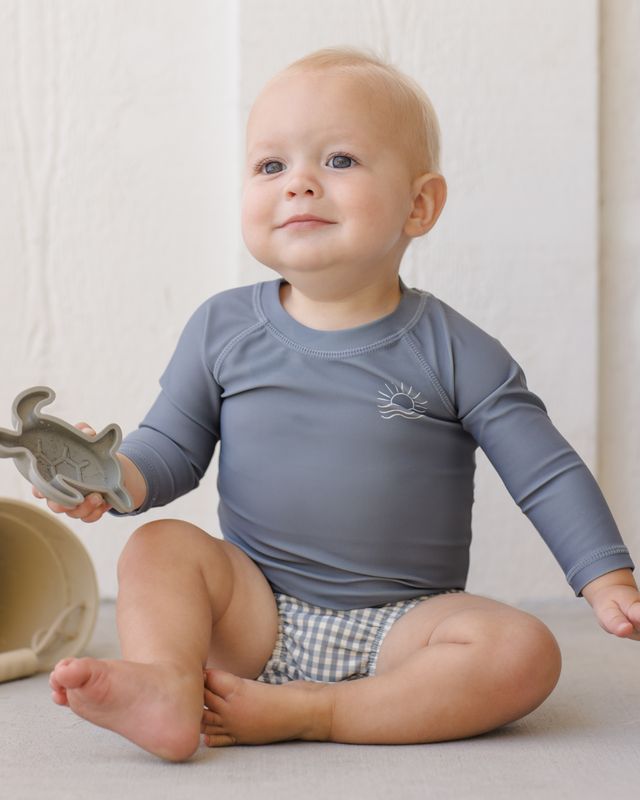 Baby wearing a blue long-sleeve shirt and checkered diaper sitting on a light-colored surface.