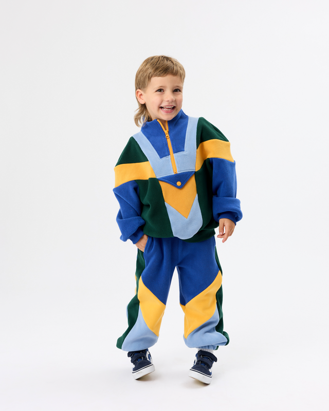 A young boy wearing a colorful color-blocked tracksuit with blue, green, yellow, and light blue panels, standing against a plain white studio background.