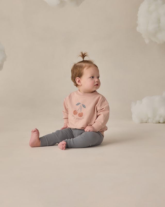Baby sitting on a beige surface wearing a pink sweatshirt with a cherry design and gray pants.