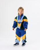 A young boy modeling a matching two-piece color-block fleece tracksuit with geometric panels in blue, yellow, green, and light blue, paired with navy sneakers, against a plain white studio background.