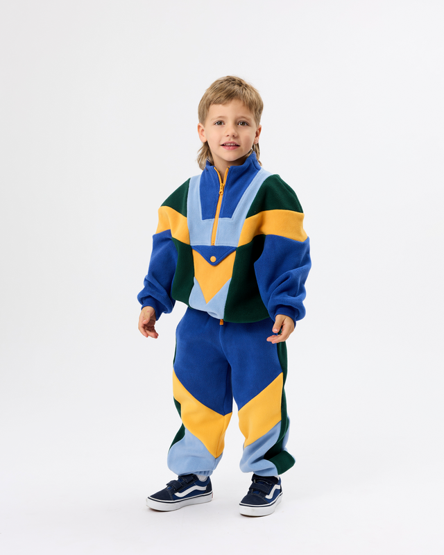 A young boy modeling a matching two-piece color-block fleece tracksuit with geometric panels in blue, yellow, green, and light blue, paired with navy sneakers, against a plain white studio background.