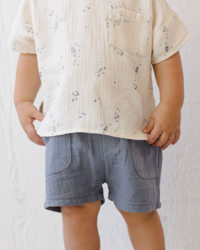 Child wearing a beige shirt with a subtle pattern and blue shorts on a plain background