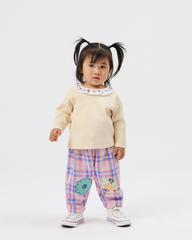 A toddler girl with black pigtails stands against a light gray studio background wearing a cream long-sleeve top with a blue patterned collar, pink and purple plaid pants with embroidered flowers, and white sneakers.