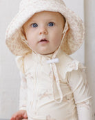 Baby wearing a cream-colored outfit with a matching hat against a light background