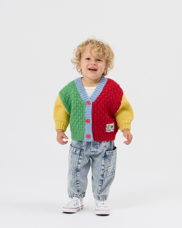 A toddler with curly blonde hair stands against a light gray studio background wearing a colorful cable-knit cardigan with green, red, yellow, and light blue sections, paired with light-wash jeans and white sneakers.
