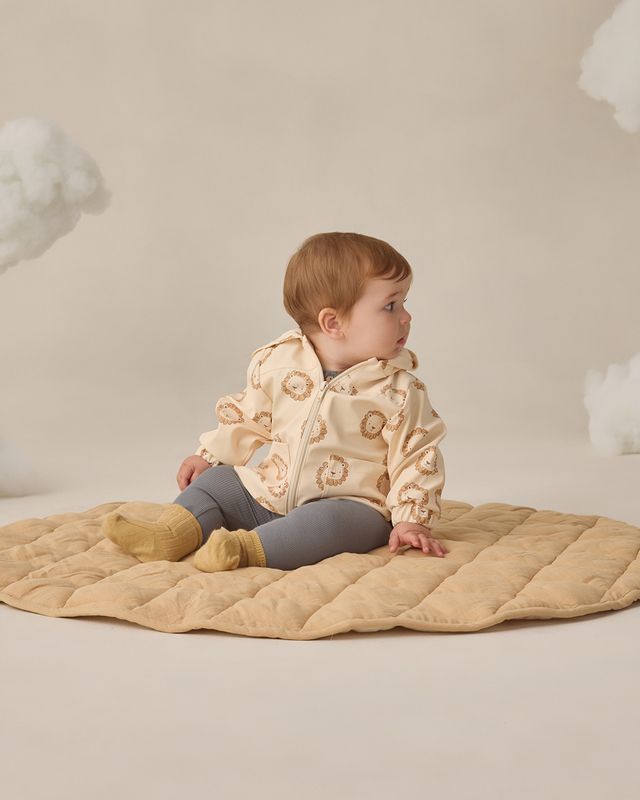 Baby sitting on a beige quilted blanket wearing a cream jacket with floral patterns against a neutral background.