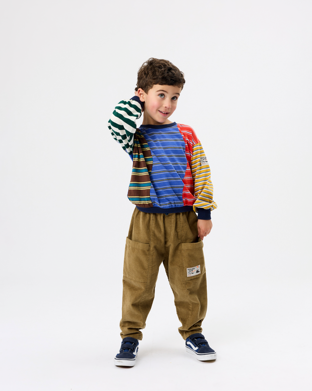 A young boy modeling a colorful patchwork sweatshirt with green, blue, red, and yellow striped panels, paired with brown corduroy pants and navy sneakers, against a plain white studio background.
