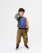 A young boy modeling a colorful patchwork sweatshirt with green, blue, red, and yellow striped panels, paired with brown corduroy pants and navy sneakers, against a plain white studio background.