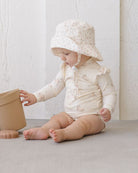 Baby in a white outfit and floral hat playing with a beige pot on a light surface.