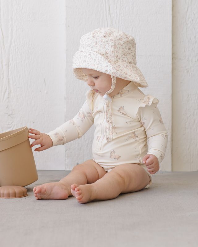 Baby in a white outfit and floral hat playing with a beige pot on a light surface.
