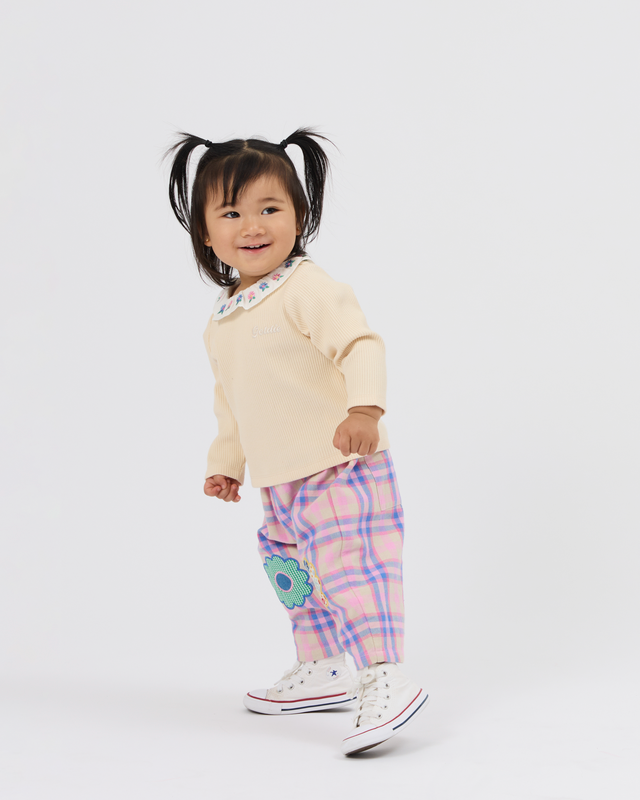 A toddler girl wearing a cream long-sleeve top, pink and purple plaid pants with a blue flower applique, and white canvas sneakers, standing against a light gray studio background.