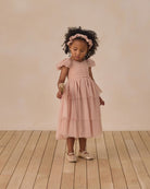 Young girl in a pink dress standing on a wooden floor with a plain background