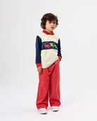 A child model wearing a color-blocked long-sleeve polo shirt with a colorful graphic across the chest and matching red wide-leg trousers, standing against a plain white studio background.