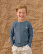 Child wearing a blue long-sleeve shirt with a logo against a brown textured background