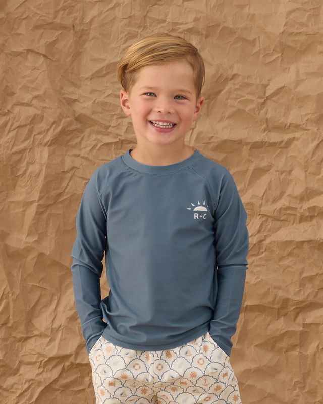Child wearing a blue long-sleeve shirt with a logo against a brown textured background