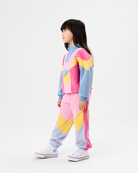 A child model wearing a pastel color-blocked tracksuit with pink, light blue, and yellow geometric panels, paired with white sneakers, photographed against a seamless white studio background.