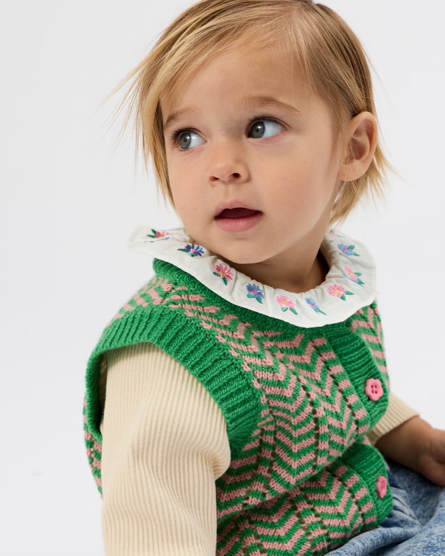 A close-up of a young child wearing a green and pink knit vest over a cream ribbed long-sleeve top, with a white floral collar, against a plain light gray background.
