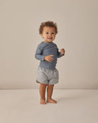Child wearing a blue long-sleeve shirt and checkered shorts on a beige background