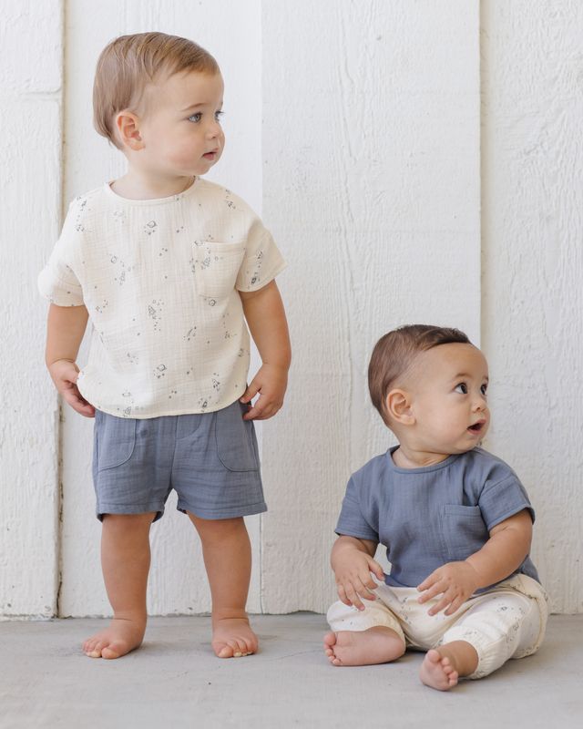 Two toddlers standing against a white wall, one wearing a white shirt with small patterns and blue shorts, the other in a blue shirt and beige pants.