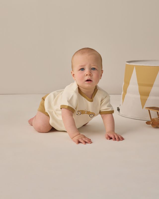 Baby in a white and brown onesie sitting on a light-colored floor with a neutral background