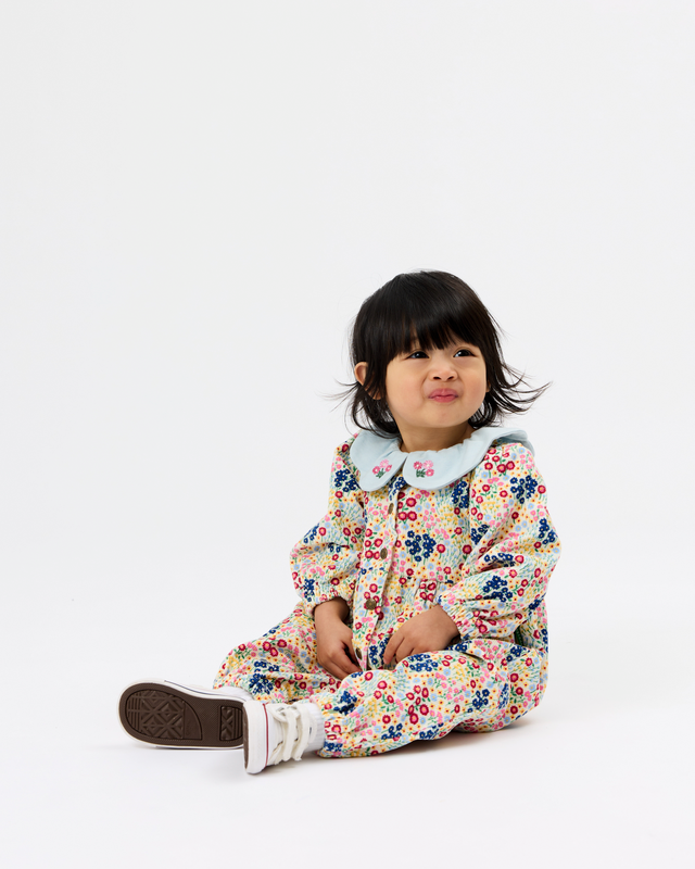 A toddler sitting on a white studio floor wearing a long-sleeve floral jumpsuit with a light blue Peter Pan collar and white sneakers.