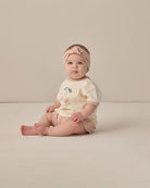 Baby sitting on a plain background wearing a light-colored outfit with a headband.