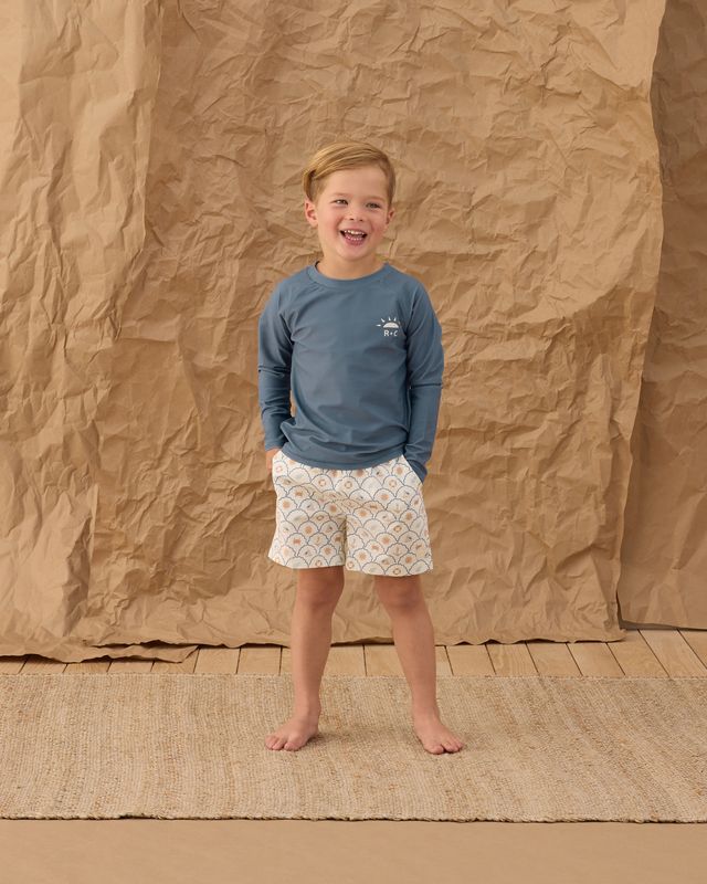 Child wearing a blue long-sleeve shirt and patterned shorts standing against a brown paper backdrop.