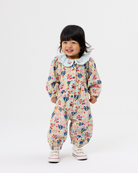 A toddler girl wearing a cream-colored floral jumpsuit with a light blue Peter Pan collar, standing against a plain white studio background.