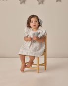 Child in a checkered dress sitting on a stool holding gray teddy bears against a neutral background