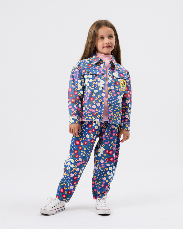 A young girl modeling a matching two-piece floral outfit consisting of a long-sleeve button-up shirt and pants, featuring a blue base with pink, red, yellow, and white flowers, paired with white sneakers on a light gray background.