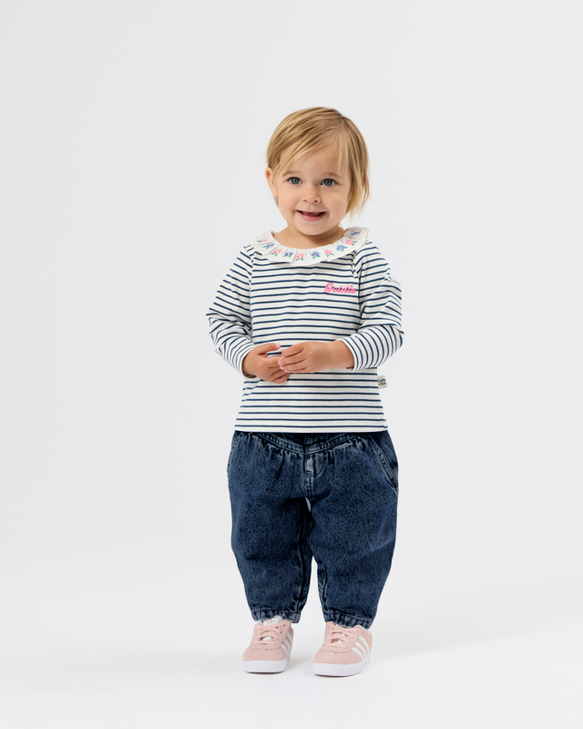 A toddler girl standing against a white studio background, wearing a navy and white striped long-sleeve top with a ruffled collar, dark blue denim pants, and light pink sneakers.