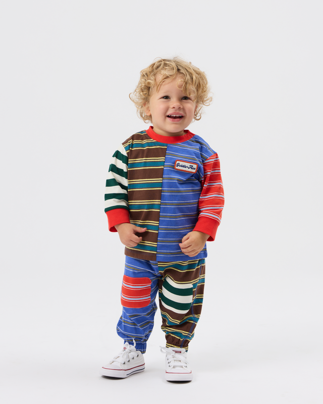 A toddler with curly blonde hair stands against a light gray studio background wearing a colorful striped one-piece jumpsuit with red, blue, brown, green, and white horizontal bands, plus white sneakers.