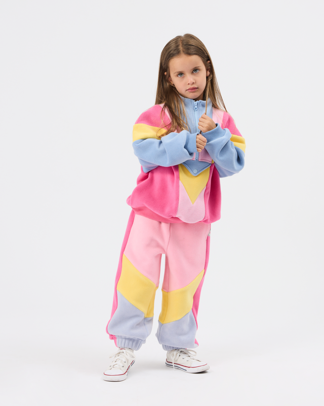 A young girl modeling a pastel color-blocked tracksuit consisting of a zip-up jacket and matching sweatpants. The outfit features blocks of pink, light blue, and yellow, paired with white sneakers, set against a plain light gray studio background.