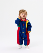 A toddler standing against a plain white studio background wearing a colorful patchwork corduroy one-piece jumpsuit with blue, red, yellow, green, and white sections, plus white sneakers.