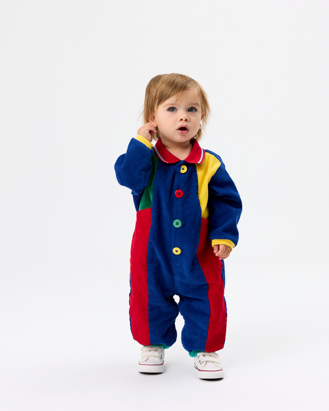 A toddler standing against a plain white studio background wearing a colorful patchwork corduroy one-piece jumpsuit with blue, red, yellow, green, and white sections, plus white sneakers.