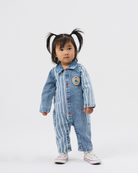 A toddler girl with dark hair in pigtails stands against a light gray studio background wearing a light blue denim one-piece jumpsuit with white vertical stripes, red buttons, and a circular patch on the chest, paired with white sneakers.