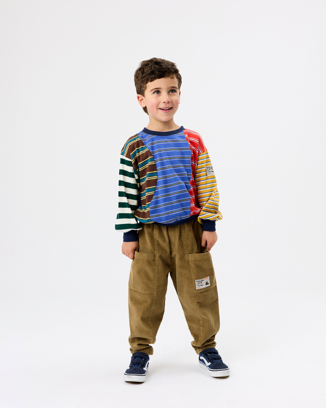 A young boy modeling a colorful long-sleeve knit top with striped and solid color panels, paired with brown pants and navy sneakers, against a plain white studio background.