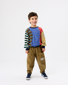 A young boy modeling a colorful long-sleeve knit top with striped and solid color panels, paired with brown pants and navy sneakers, against a plain white studio background.