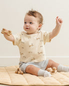 Baby wearing a cream-colored shirt with small patterns, sitting on a beige blanket.