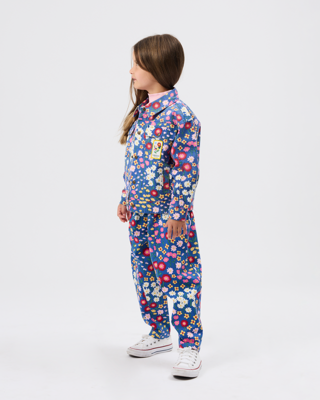 A child model shown in profile wearing a blue one-piece tracksuit with a dense multicolor floral print, paired with white sneakers, against a plain light gray studio background.