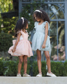 Two young girls in dresses standing outdoors with greenery in the background