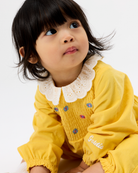A young child wearing a yellow long-sleeve top with a white eyelet Peter Pan collar and small embroidered floral details, photographed against a plain light background.