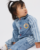 A toddler girl wearing a light blue denim one-piece jumpsuit with white vertical stripes, red buttons, and a cartoon bear patch on the chest, sitting against a plain white background.