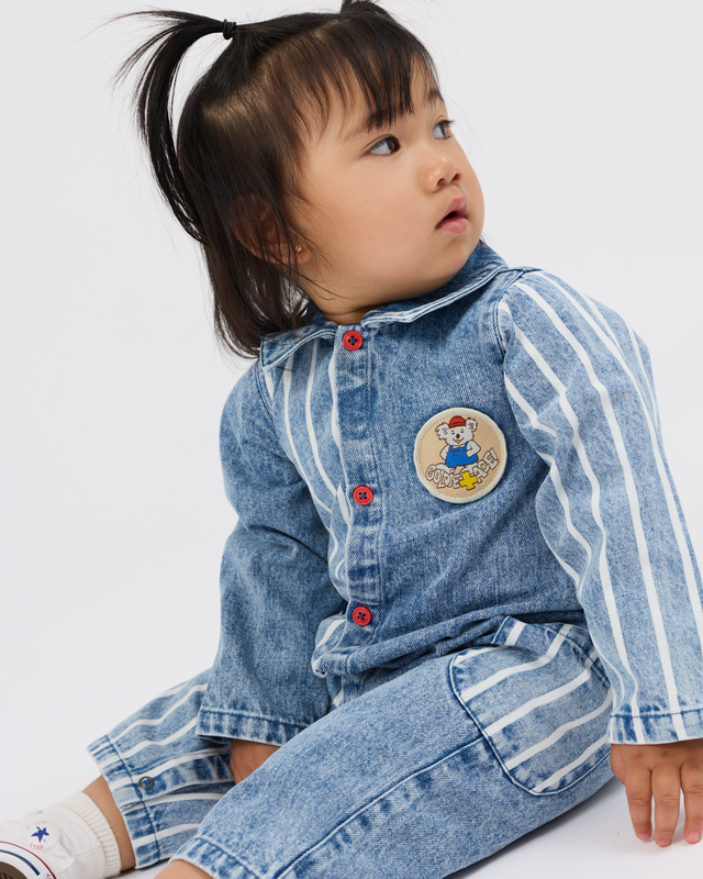 A toddler girl wearing a light blue denim one-piece jumpsuit with white vertical stripes, red buttons, and a cartoon bear patch on the chest, sitting against a plain white background.