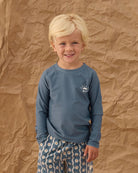 Young boy wearing a blue long-sleeve shirt and patterned pants standing against a brown textured background