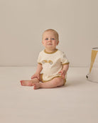 Baby wearing a white and brown outfit with a car design, sitting on a beige surface.