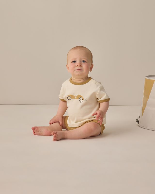 Baby wearing a white and brown outfit with a car design, sitting on a beige surface.
