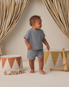 Baby in a blue outfit standing in a room with decorative curtains and toys.